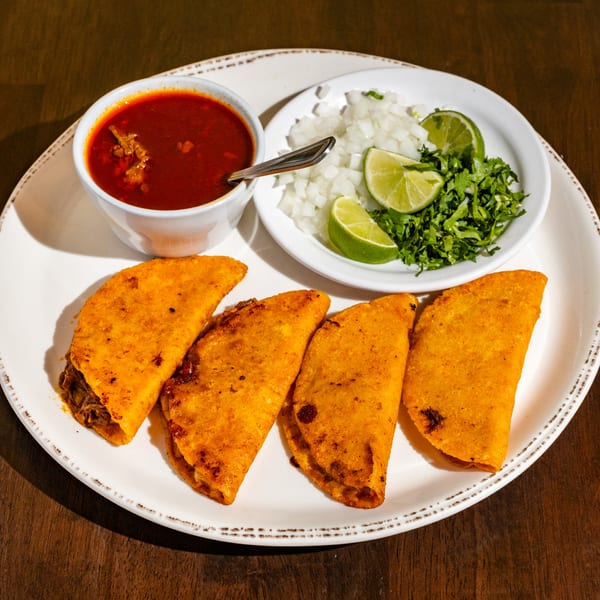 Four Chuyita's birria tacos with a side of consommé, diced onions, cilantro, and lime wedges.