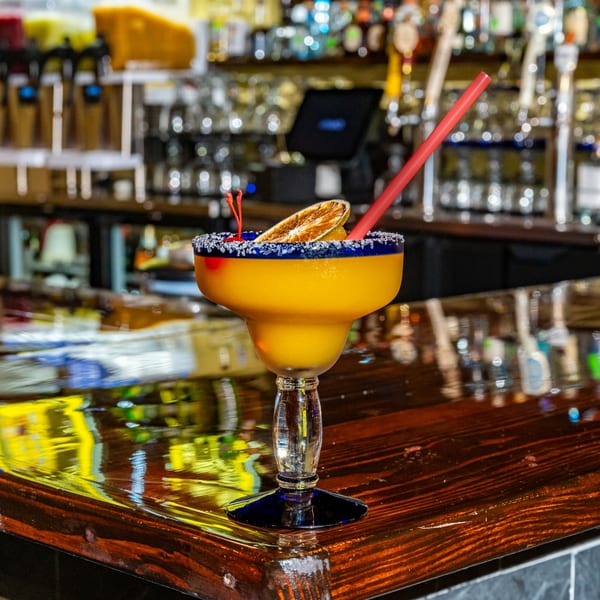 A vibrant yellow margarita with a salted rim, cherry, and dried orange slice sits on a bar.