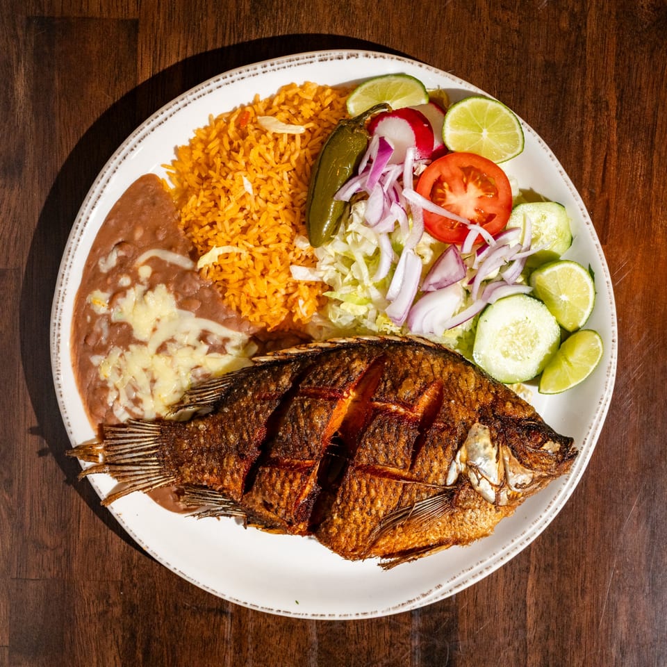 Fried whole fish with rice, refried beans, and a side salad with lime on a white plate.
