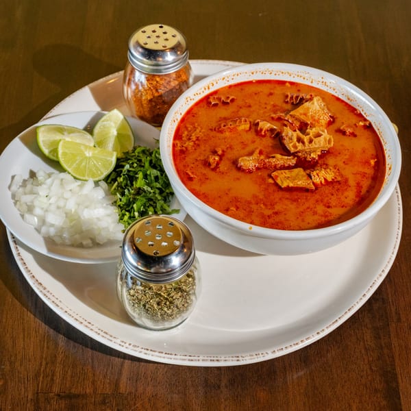 Menudo soup with tripe, red broth, and sides of diced onions, cilantro, and lime wedges.